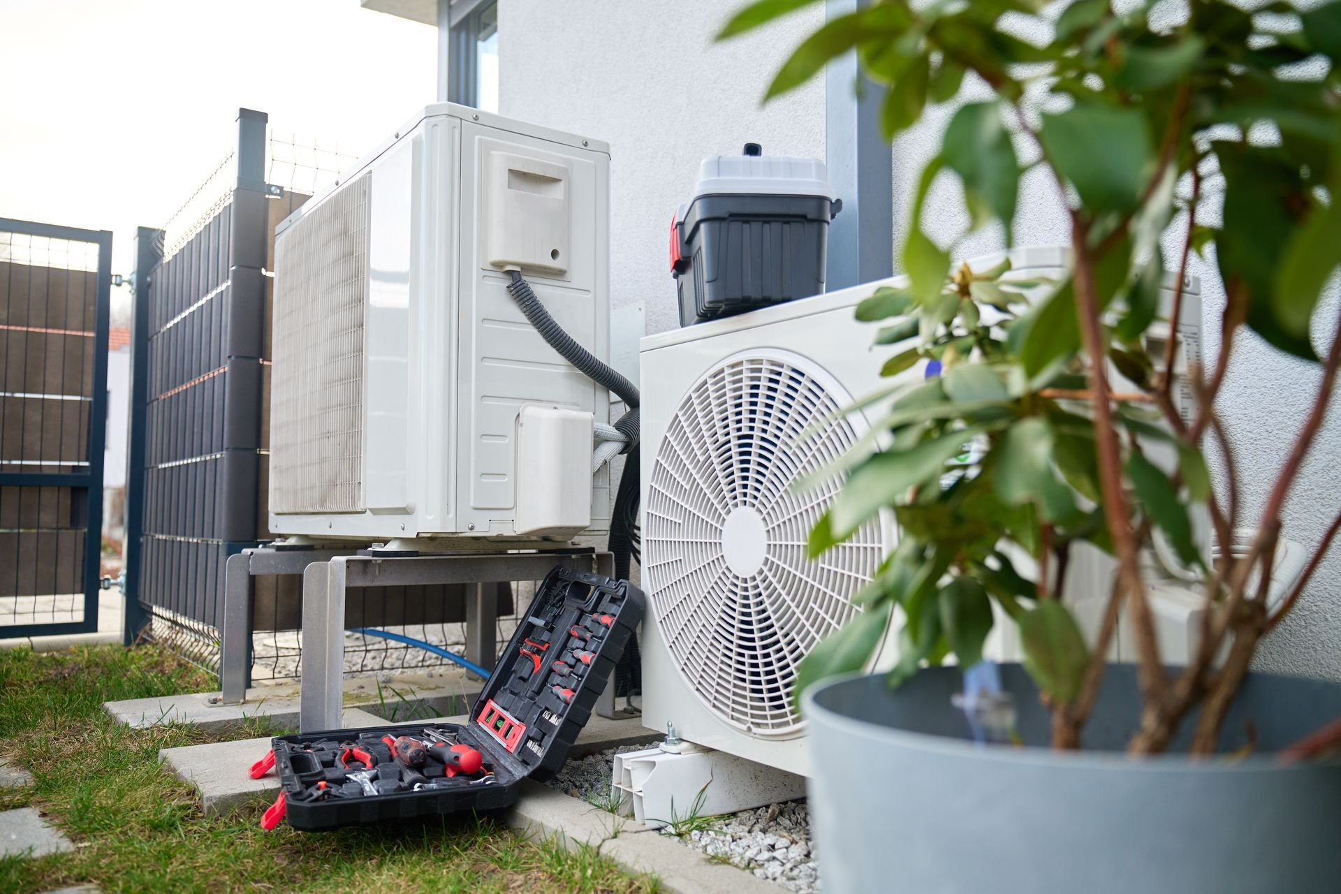 Outdoor HVAC units beside a fence, with a plant pot and tools on the grass.