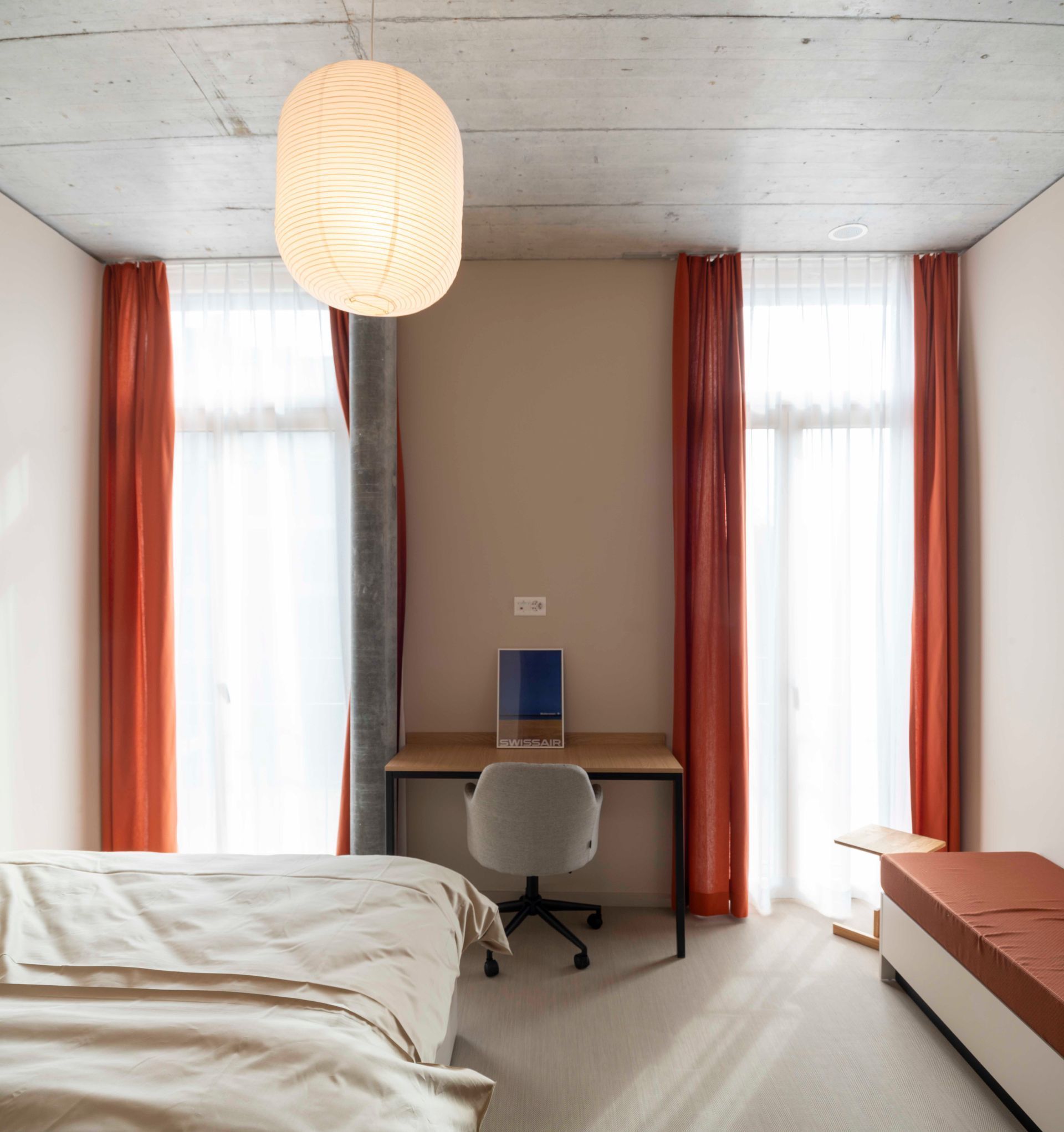 Minimal bedroom with twin windows, orange curtains, desk, chair, bed, and pendant light