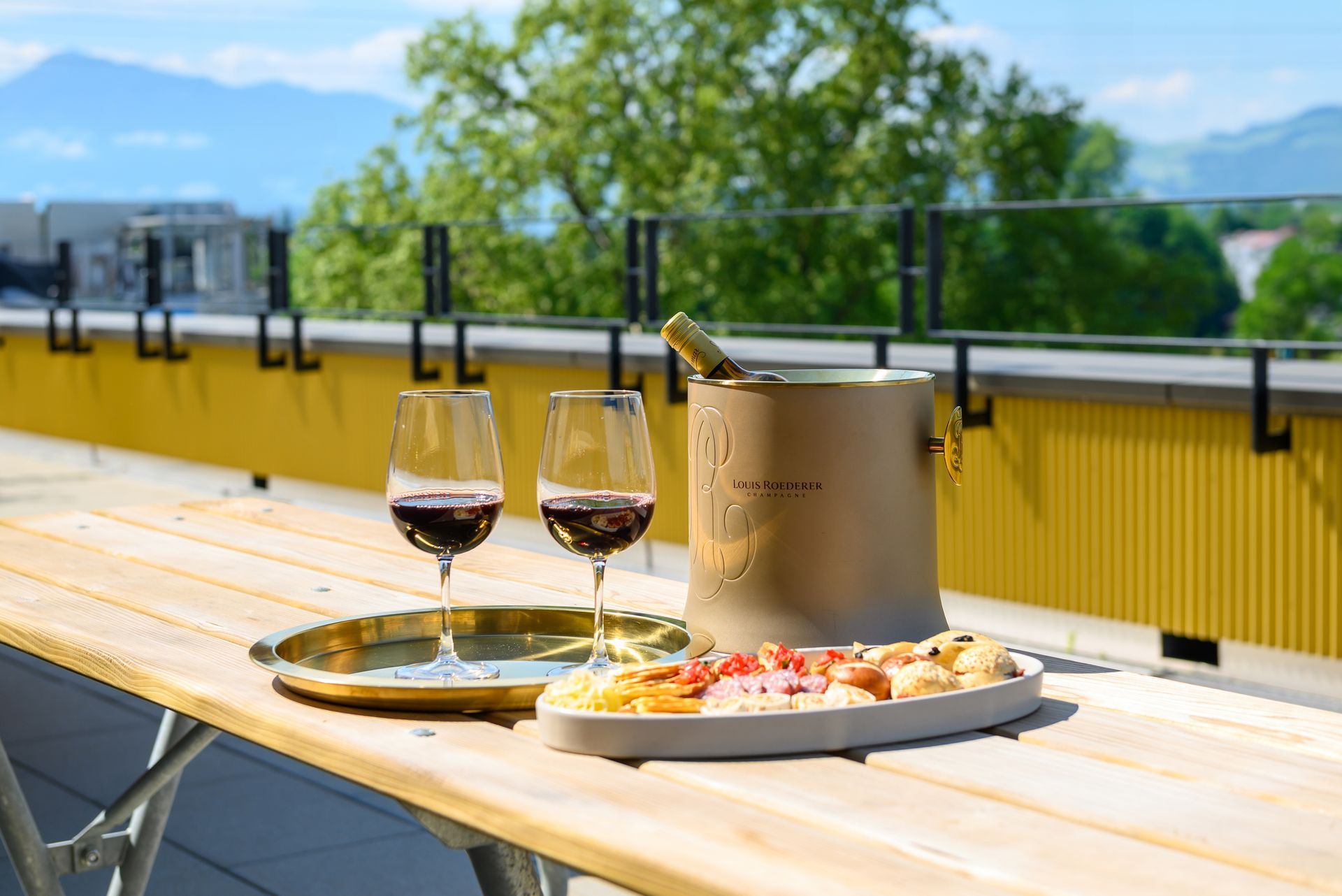 Wine glasses and a pizza on a sunlit outdoor table with mountain views in the background