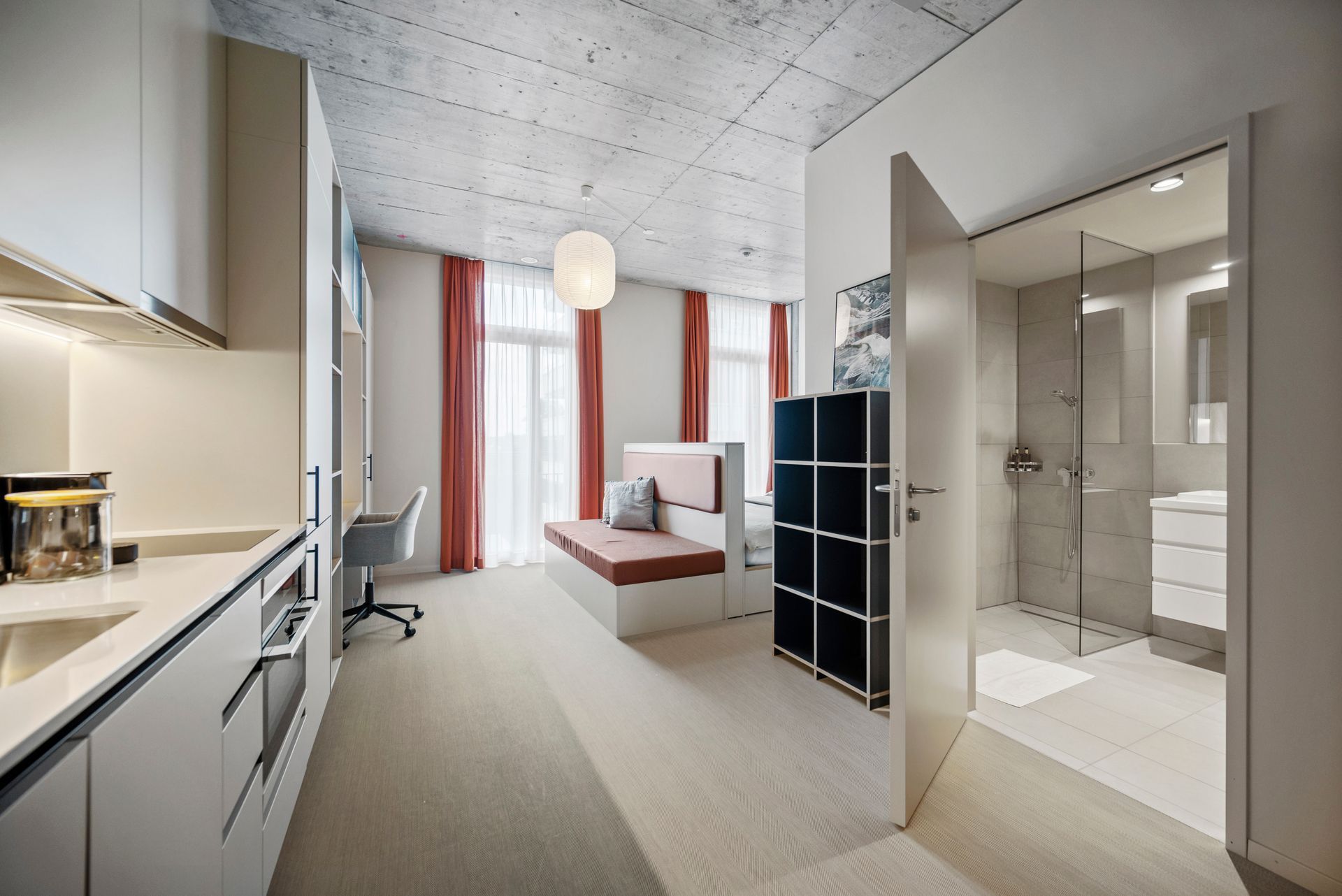 Modern studio apartment with bed, kitchenette, desk, and glass-walled bathroom