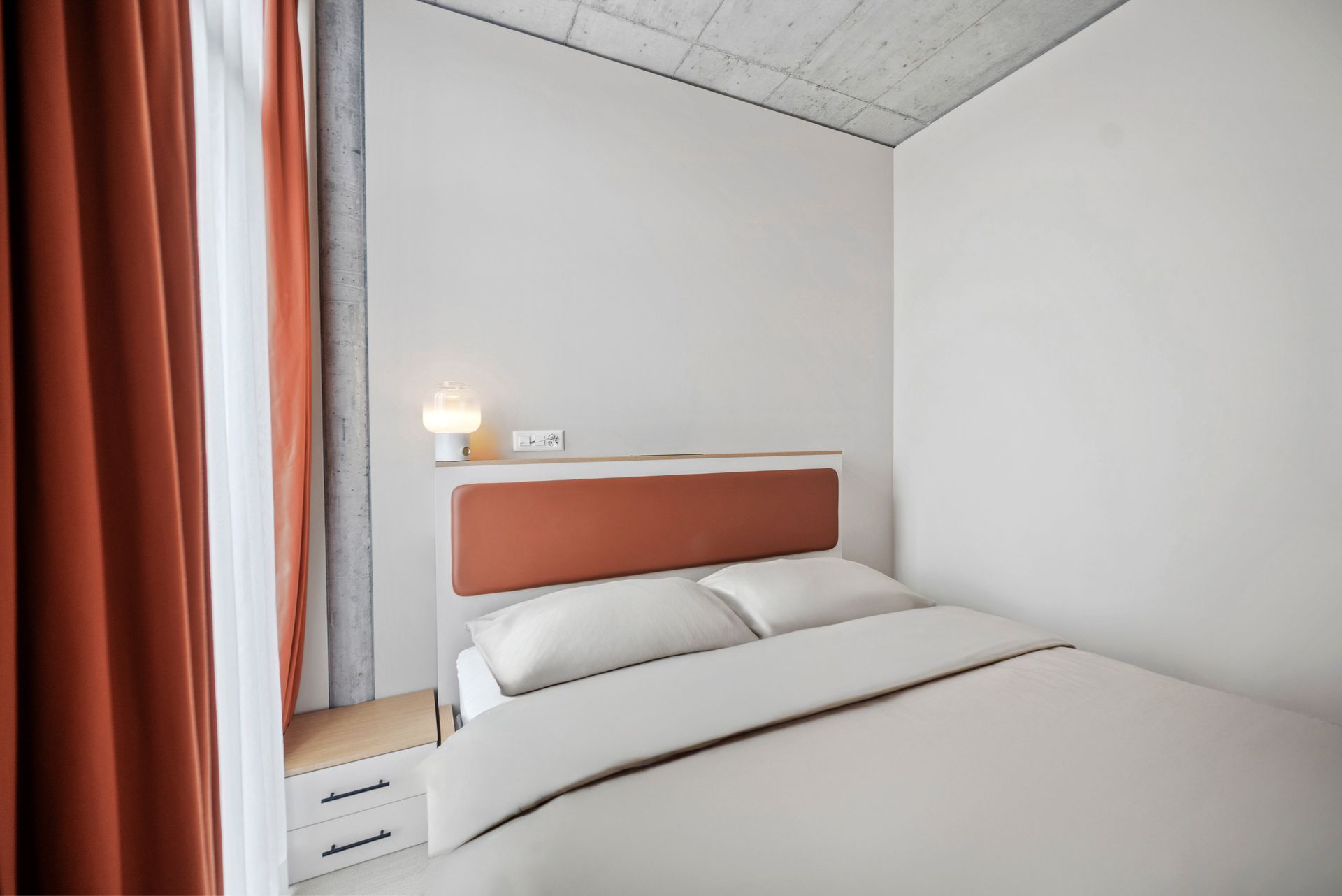 Minimal bedroom with white bedding, coral headboard, bedside lamp, and orange curtains