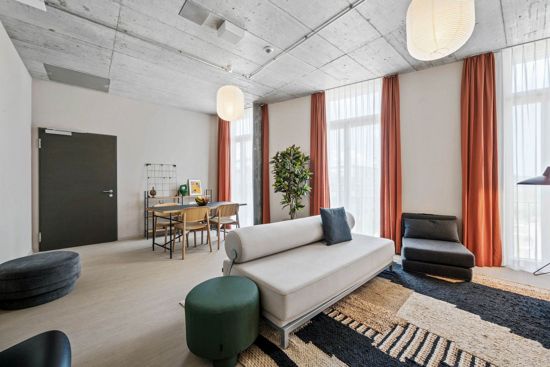 Modern living room with sofa, chairs, round ottoman, black rug, and tall windows with orange curtains