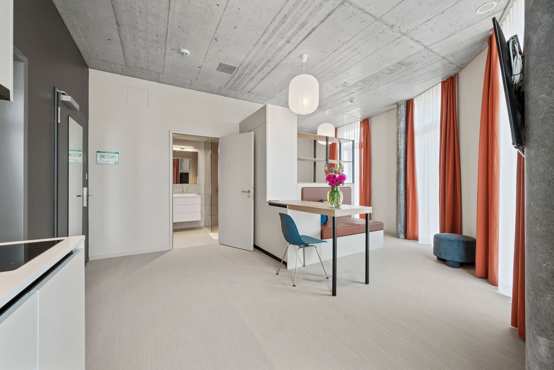 Modern apartment living room with white walls, gray carpet, orange curtains, and a small dining table near large windows