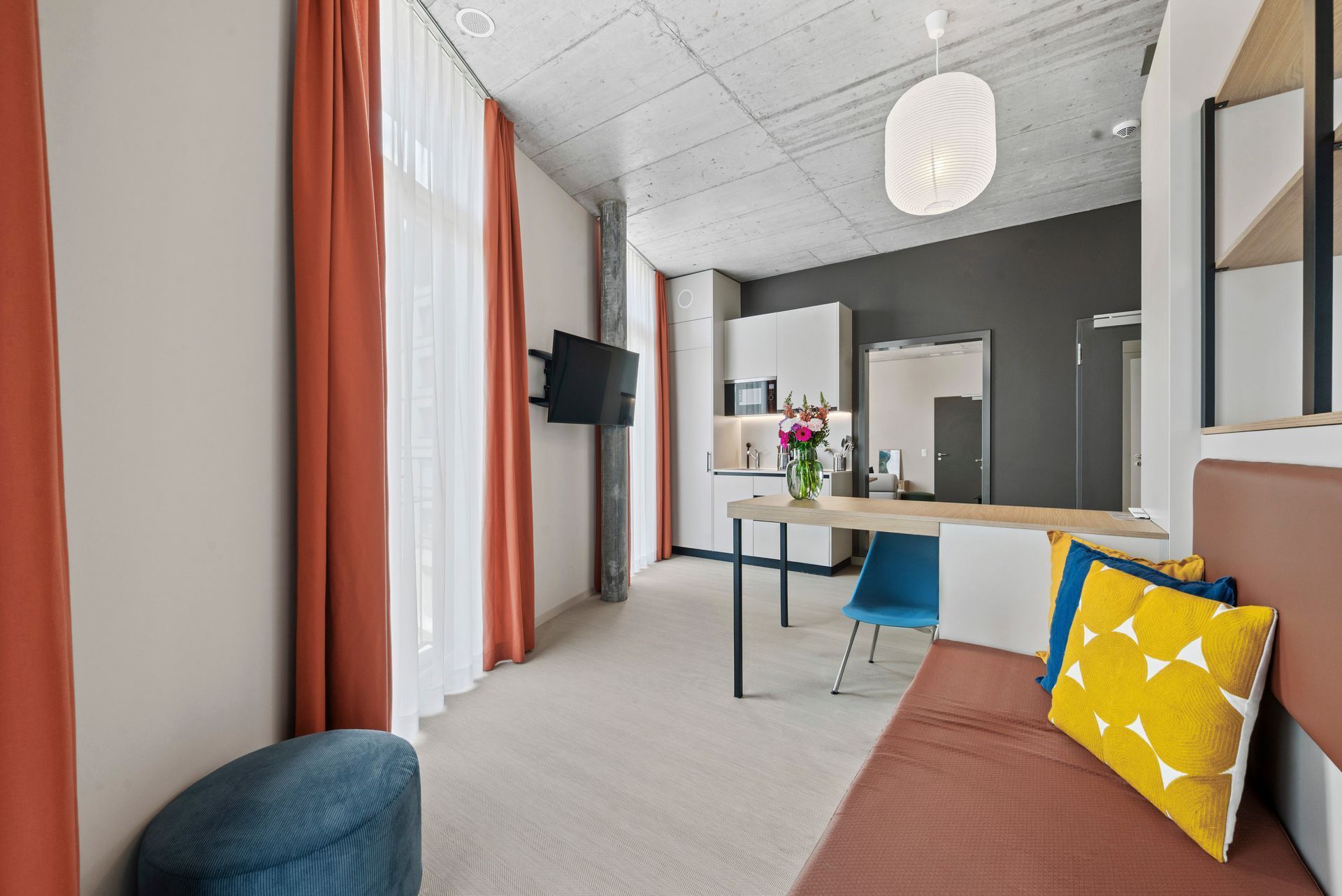 Modern living room with orange curtains, gray walls, brown sofa, yellow pillow, and a small dining area at the back