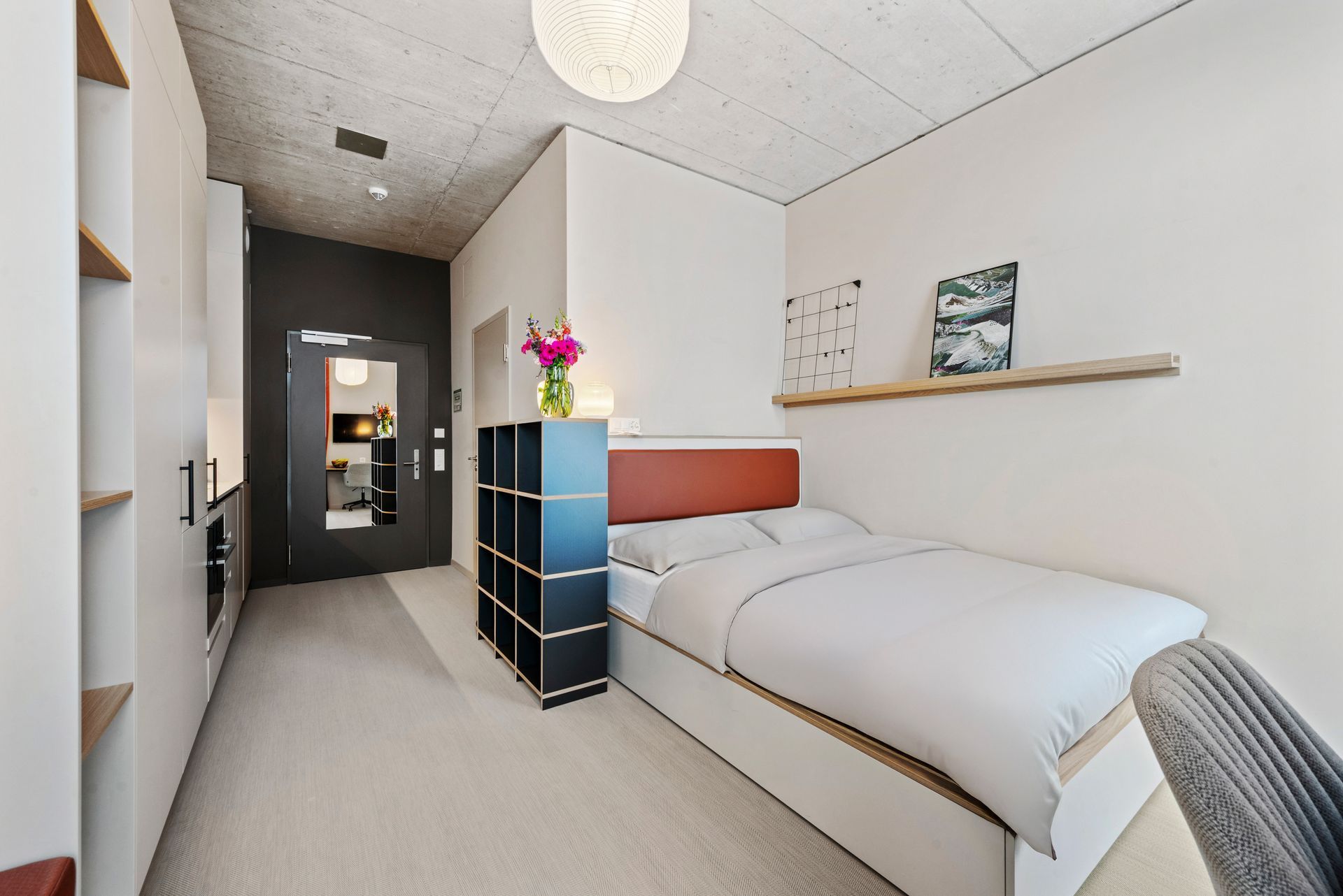 Bright minimalist bedroom with a single bed, black dresser, wall shelf, and hallway view
