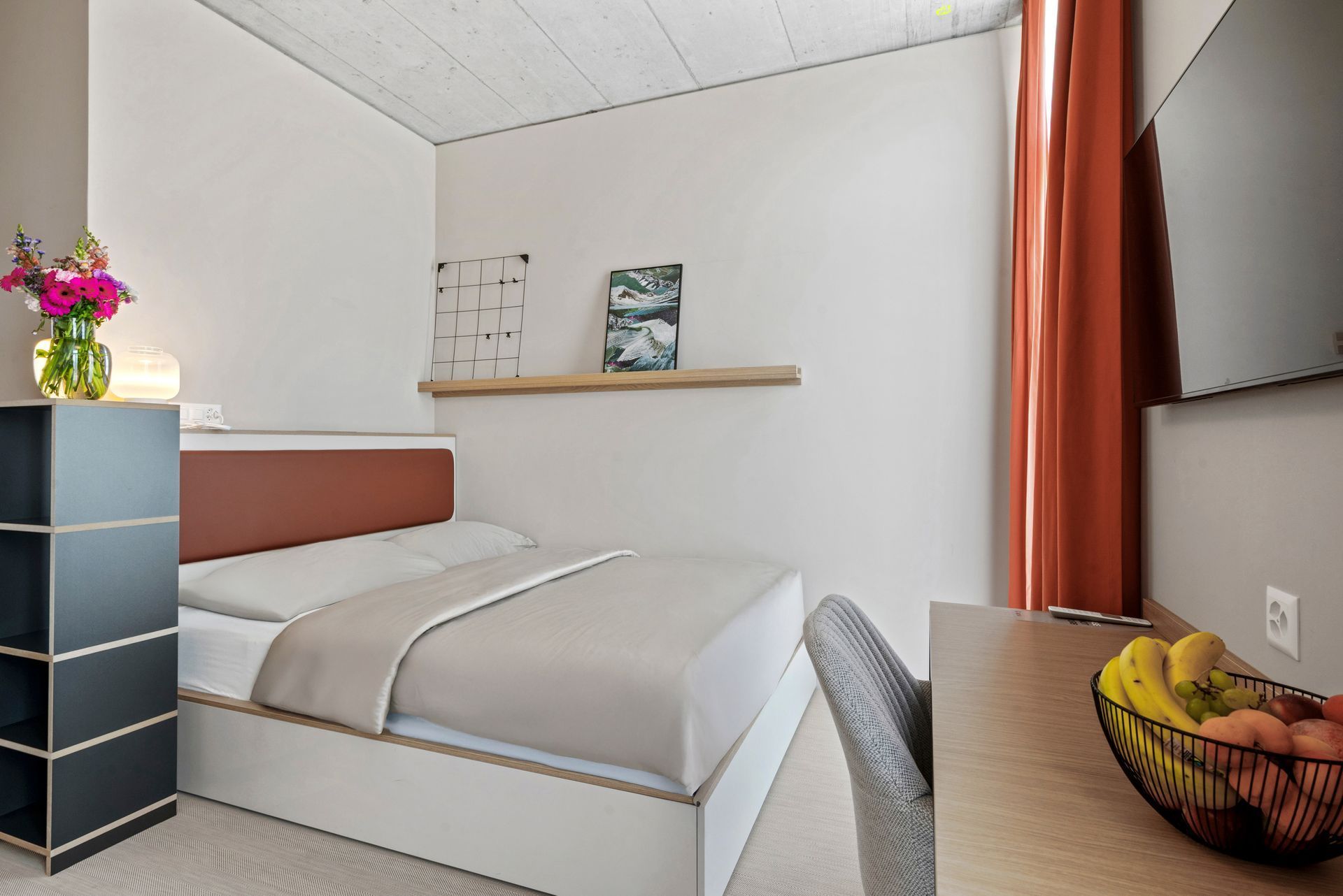 Small modern bedroom with a white bed, gray chair, wooden shelf, and a desk with fruit.