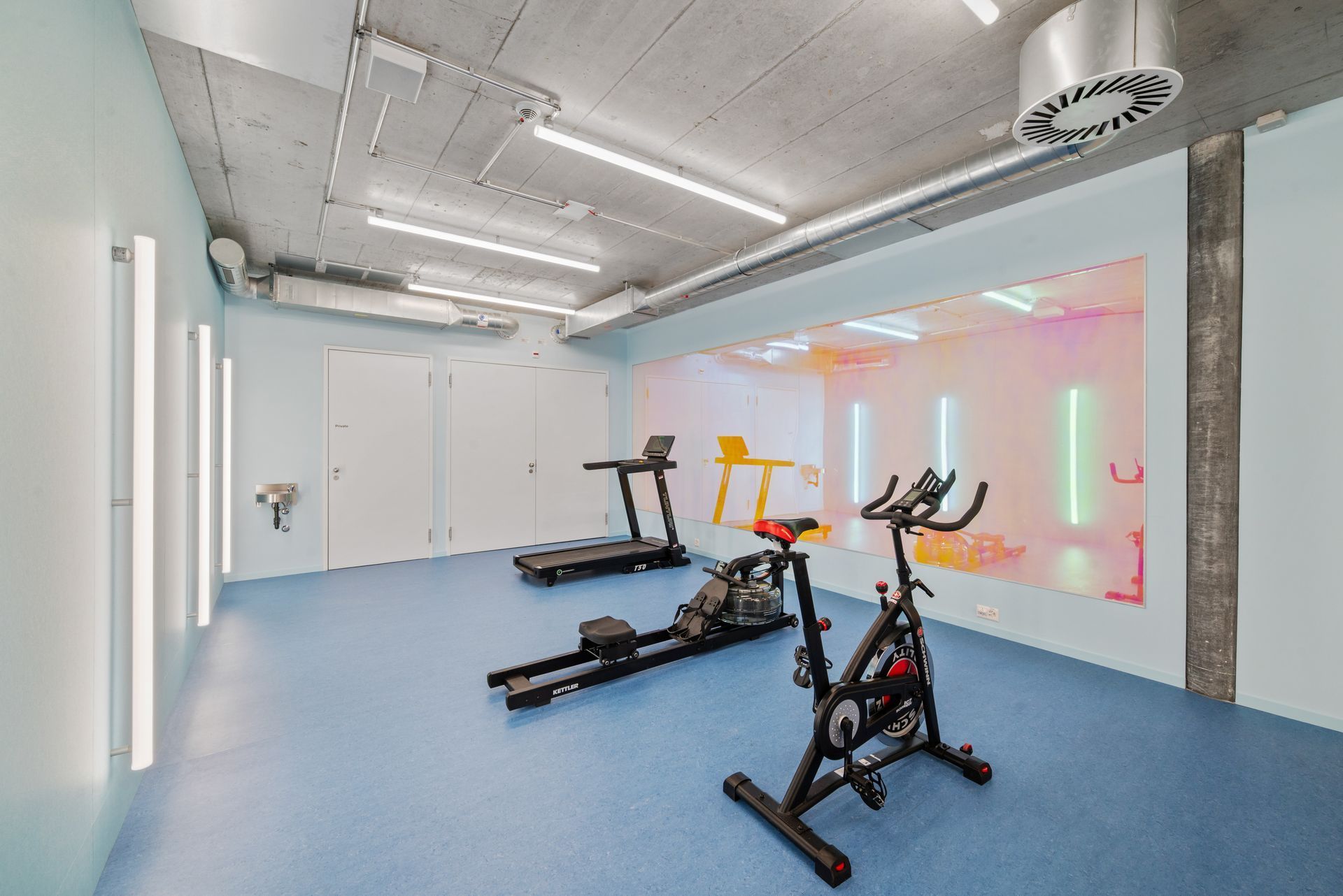 Empty gym with blue floor, white walls, and stationary exercise bikes near a mirrored wall.