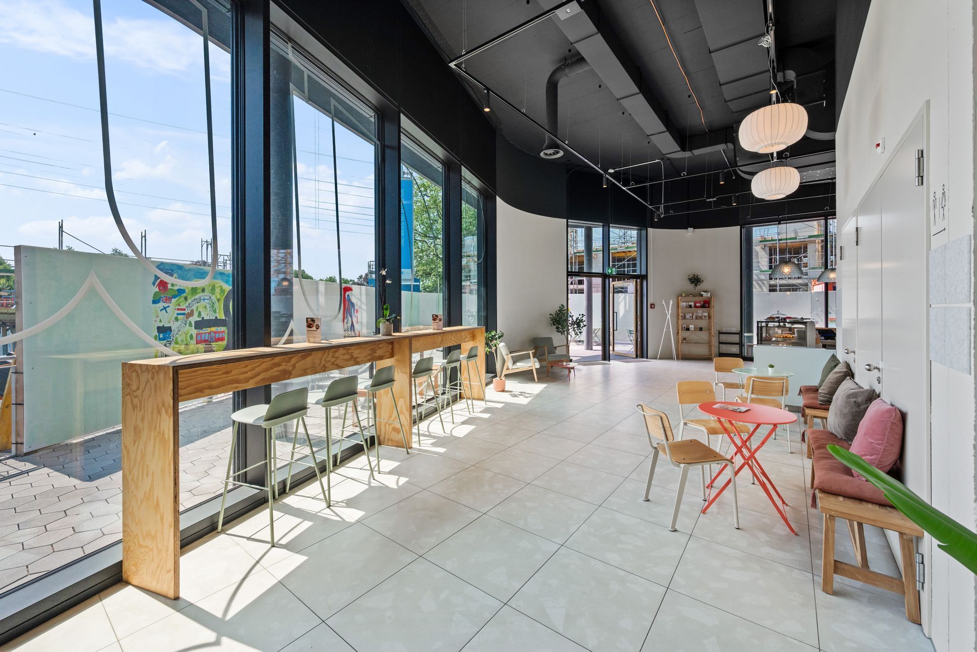 Bright modern café interior with tall windows, wooden tables, and colorful chairs by a sunlit counter.