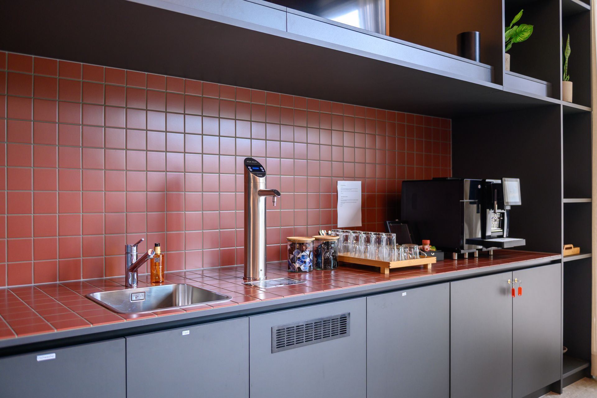 Modern kitchen counter with red tile backsplash, sink, coffee machine, and tray of cups