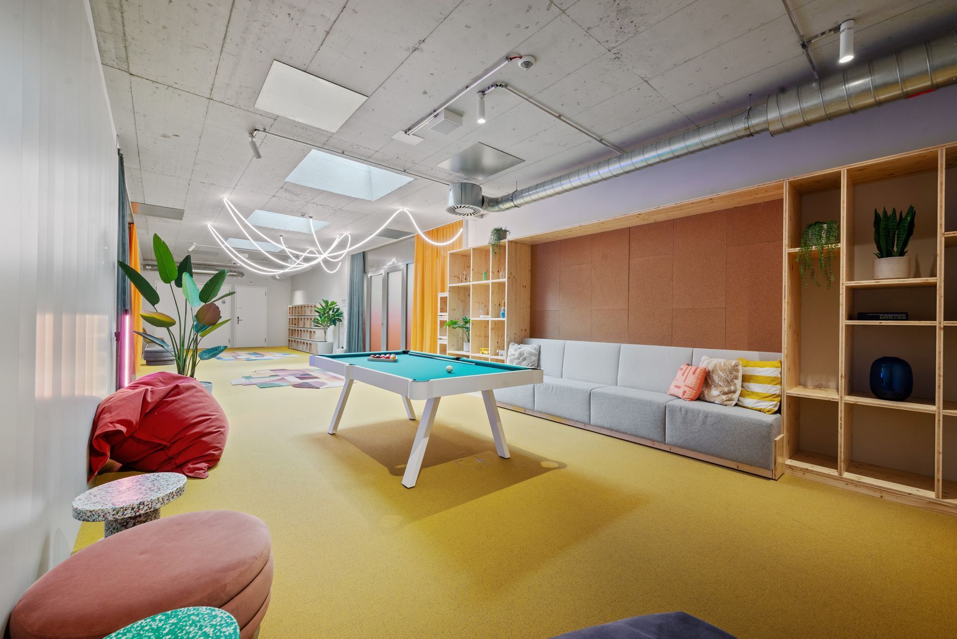 Bright modern lounge with yellow carpet, gray sofas, wooden shelves, and a foosball table.