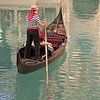 Eine Person in einer roten Jacke steht in einem kleinen Boot auf ruhigem, grünem Wasser; ein weiteres Boot liegt neben ihr.