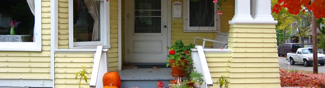 A yellow house with a porch decorated for halloween with pumpkins and flowers.