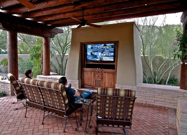 Two people are sitting on a patio under a ceiling fan watching a television.