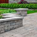 Stone garden benches beside a paved path, with green shrubs and a flower bed in the background.