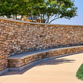 Curved stone bench beside a tan walkway and stacked-stone wall under leafy trees