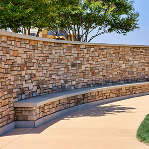 Curved stone bench beside a tan walkway and stacked-stone wall under leafy trees