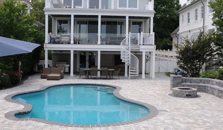 Backyard pool with stone patio and a two-story house with a white deck and stairs