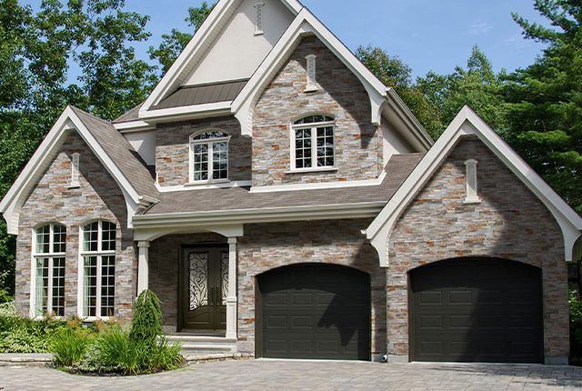 Two-story stone house with twin garage doors, arched windows, and a gabled roof surrounded by trees