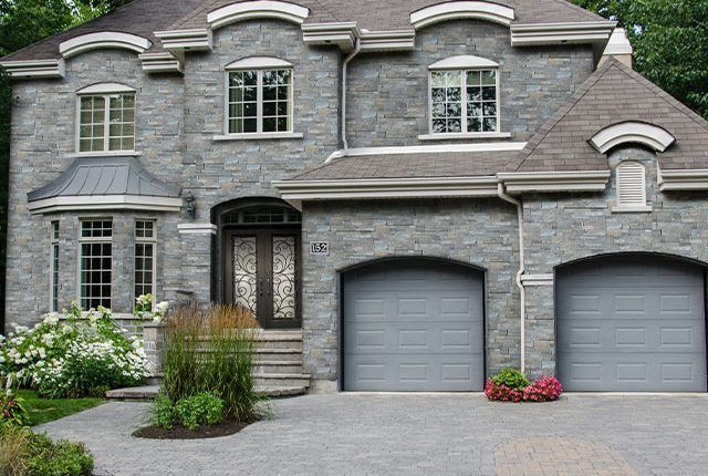 Gray stone house with arched garage doors, front steps, and landscaped garden