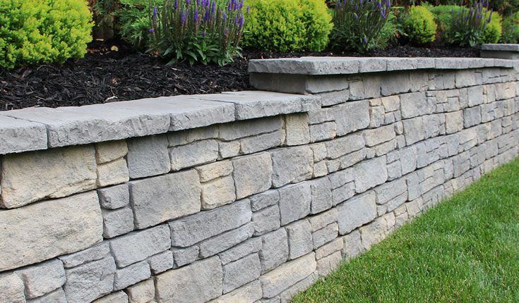 Stone retaining wall with gray stacked blocks and dark mulch beside green landscaping.