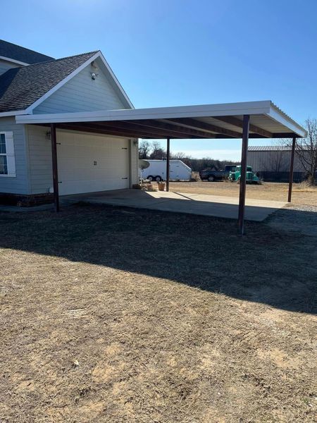 Carport attached to a light blue house, with concrete flooring and brown metal supports, under a clear sky.
