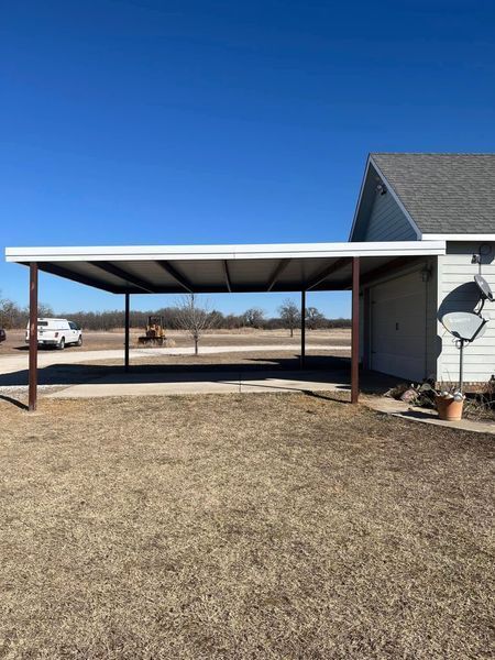 Brown-framed carport attached to a white house with a metal roof on a sunny day.