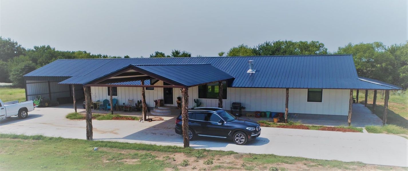 A white house with a blue roof, carport, and a black SUV on a paved driveway.