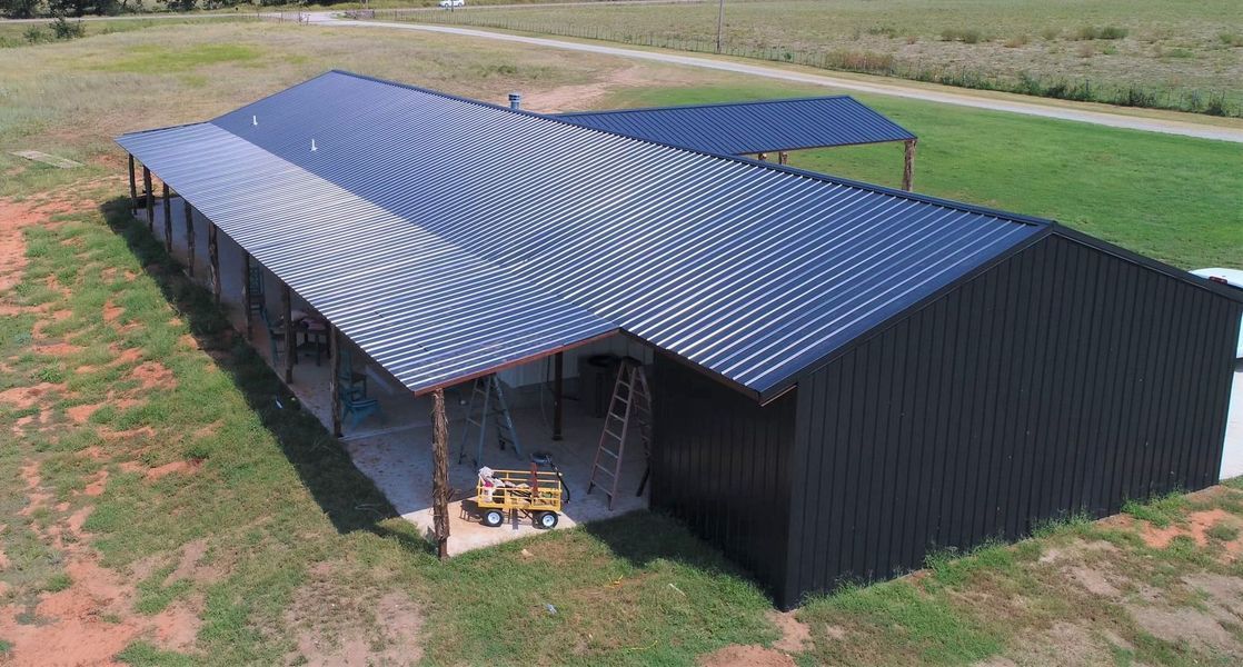 Black metal-roofed building with open-air porch, set in a grassy field on a sunny day.