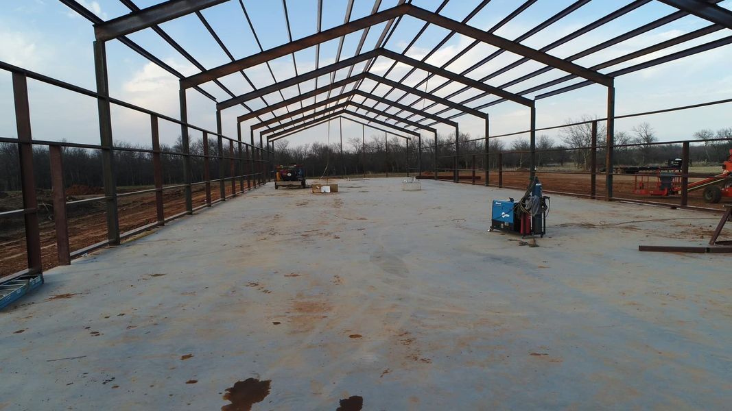 Steel frame of a building under construction on a concrete slab. Open on sides, with trees in the background.