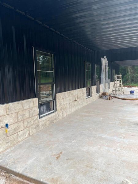 Exterior of a building under construction. Black metal siding, stone block base, concrete patio.