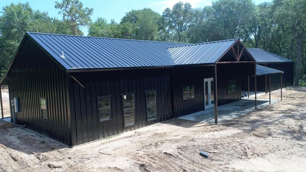 Black metal-clad building with a dark blue roof and porch, on a dirt foundation, surrounded by trees.