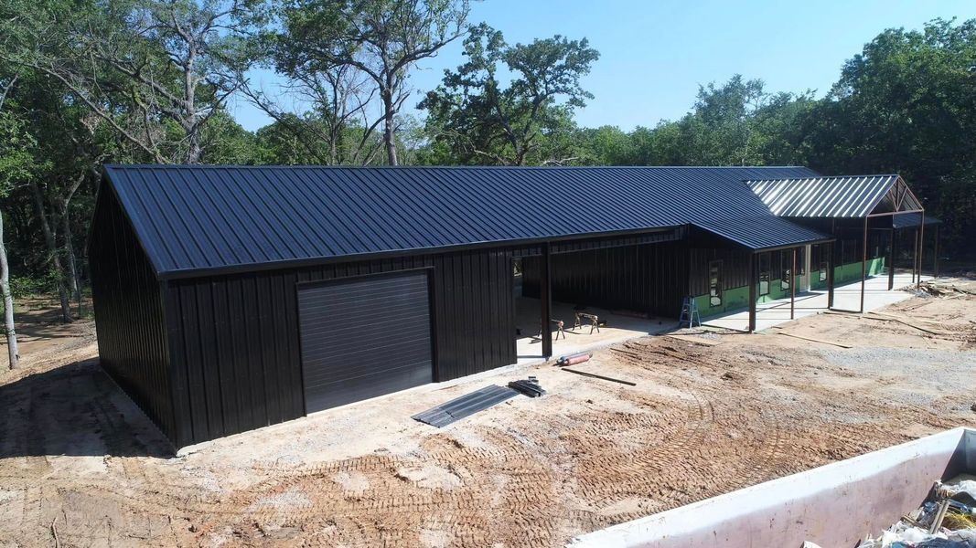 Black building with a corrugated metal roof, trees in the background, set on a sandy lot.