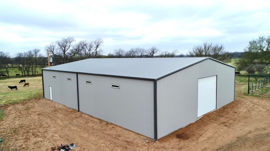 Gray metal agricultural building with a large white door, on a dirt patch, with trees and cloudy sky.