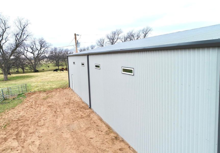 Exterior view of a long, light-colored corrugated metal building with a dirt path in the foreground and a grassy field in the background.