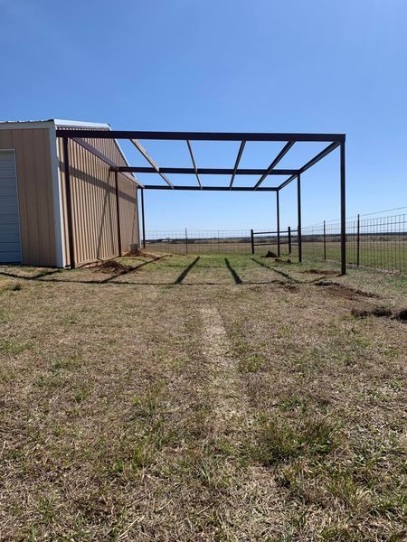 Metal frame shelter attached to a tan building with a grassy field in the foreground under a blue sky.
