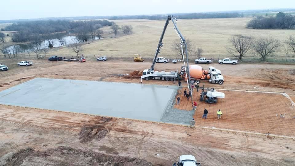 Construction site: concrete being poured. Two cement trucks with pump arms over a large rectangular area. Workers and vehicles visible.