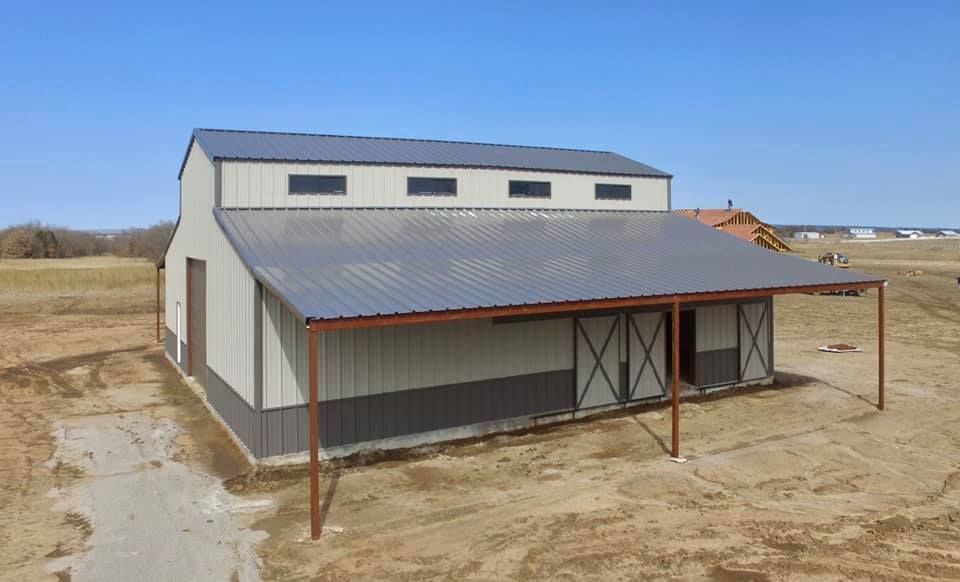Metal barn with gray and tan panels, brown roof, and an open-air awning on a sunny day.