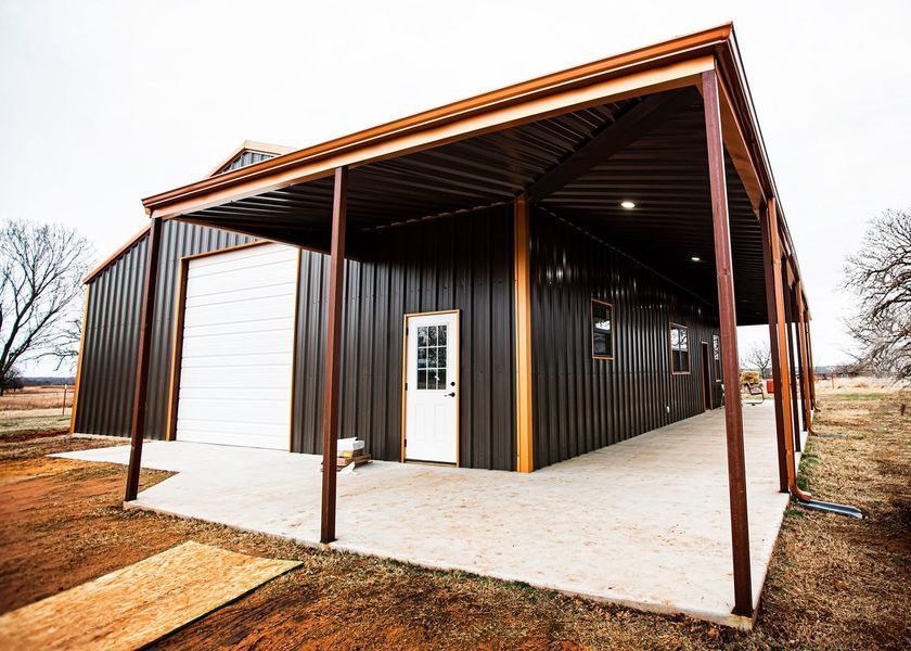 Dark brown metal building with covered porch, white door and garage door, concrete base.