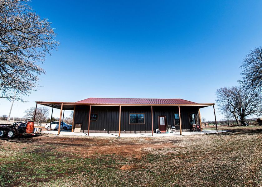 Brown metal-clad house with a red roof, porch, and clear blue sky in rural setting.