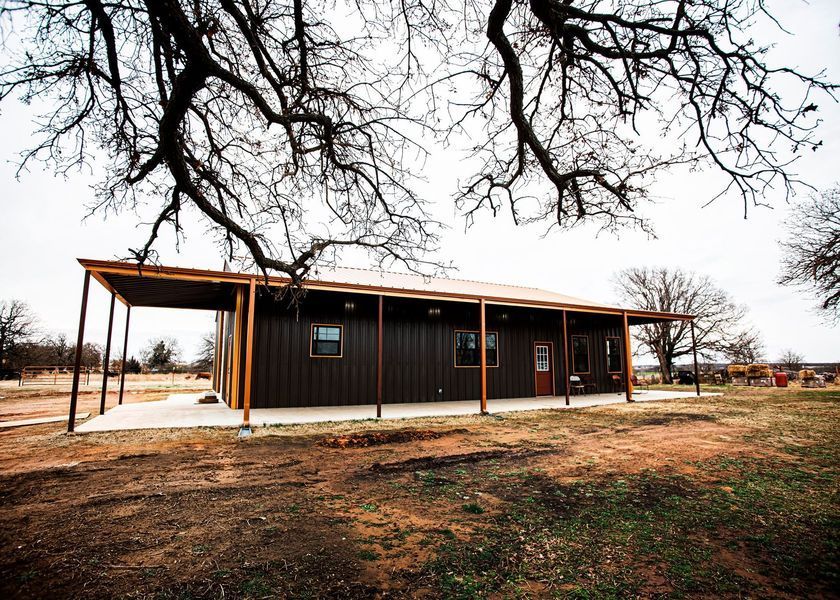 Brown metal-clad house with a porch and surrounding leafless trees under an overcast sky.