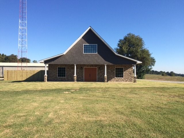 Brick barn-style house with dark roof and red doors on a grassy field under a clear blue sky