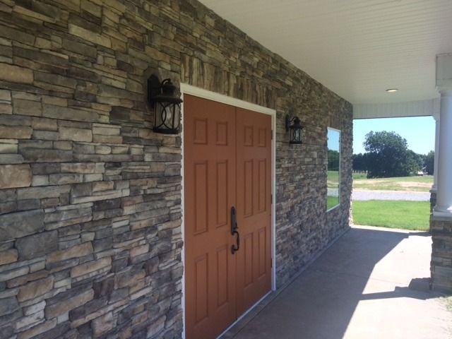 Front porch with stone walls, a brown front door, and wall lanterns in bright daylight
