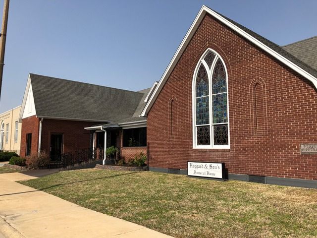 Brick church building with a tall arched window and a small sign in front on a sunny day