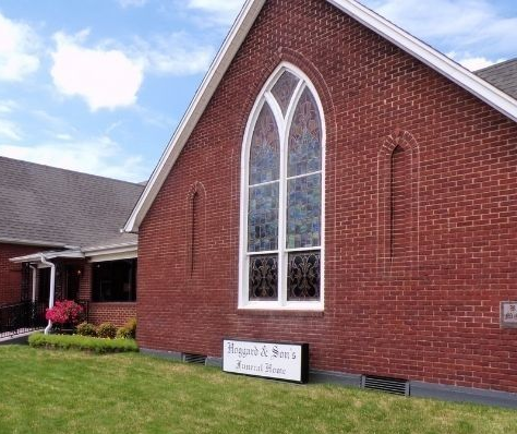 Brick church building with tall arched window and white sign in front under a blue sky