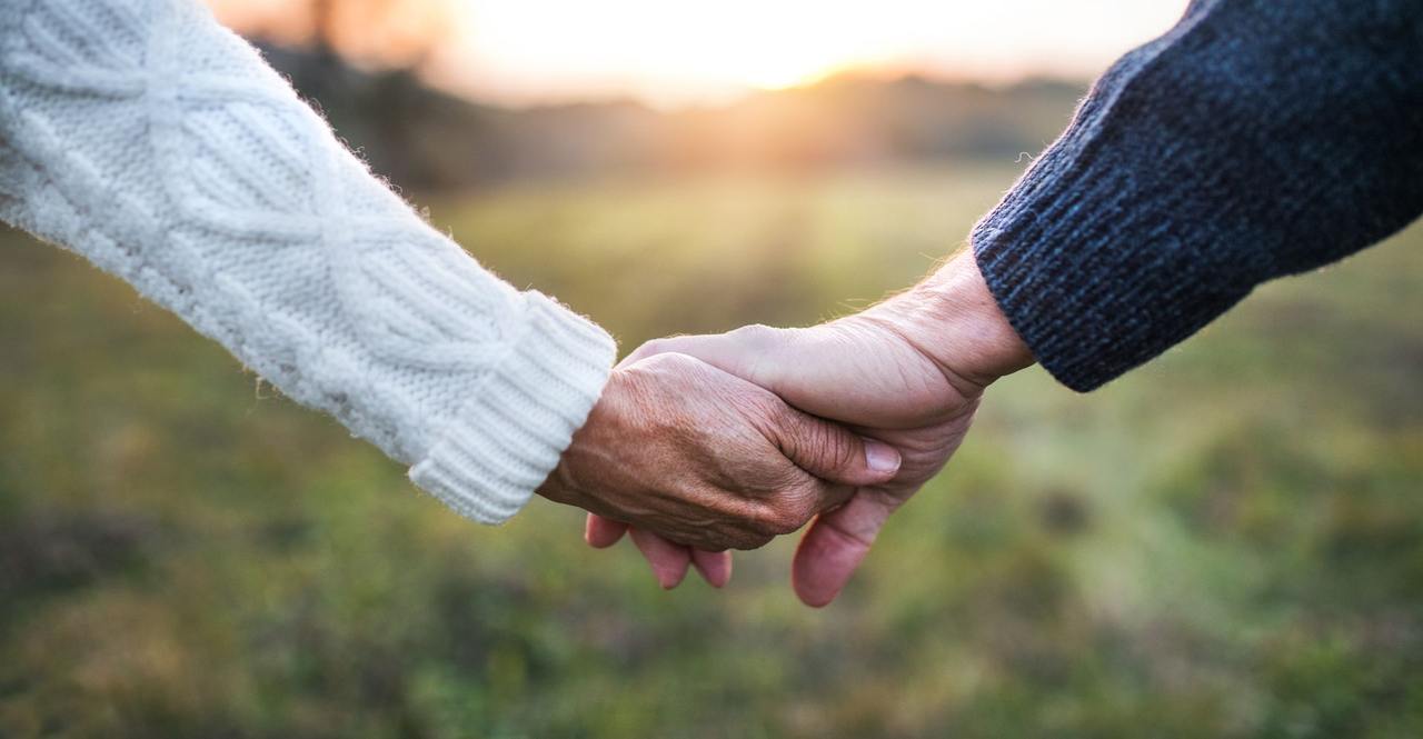 Two people holding hands outdoors at sunset, one in a white sweater and one in a blue sweater.
