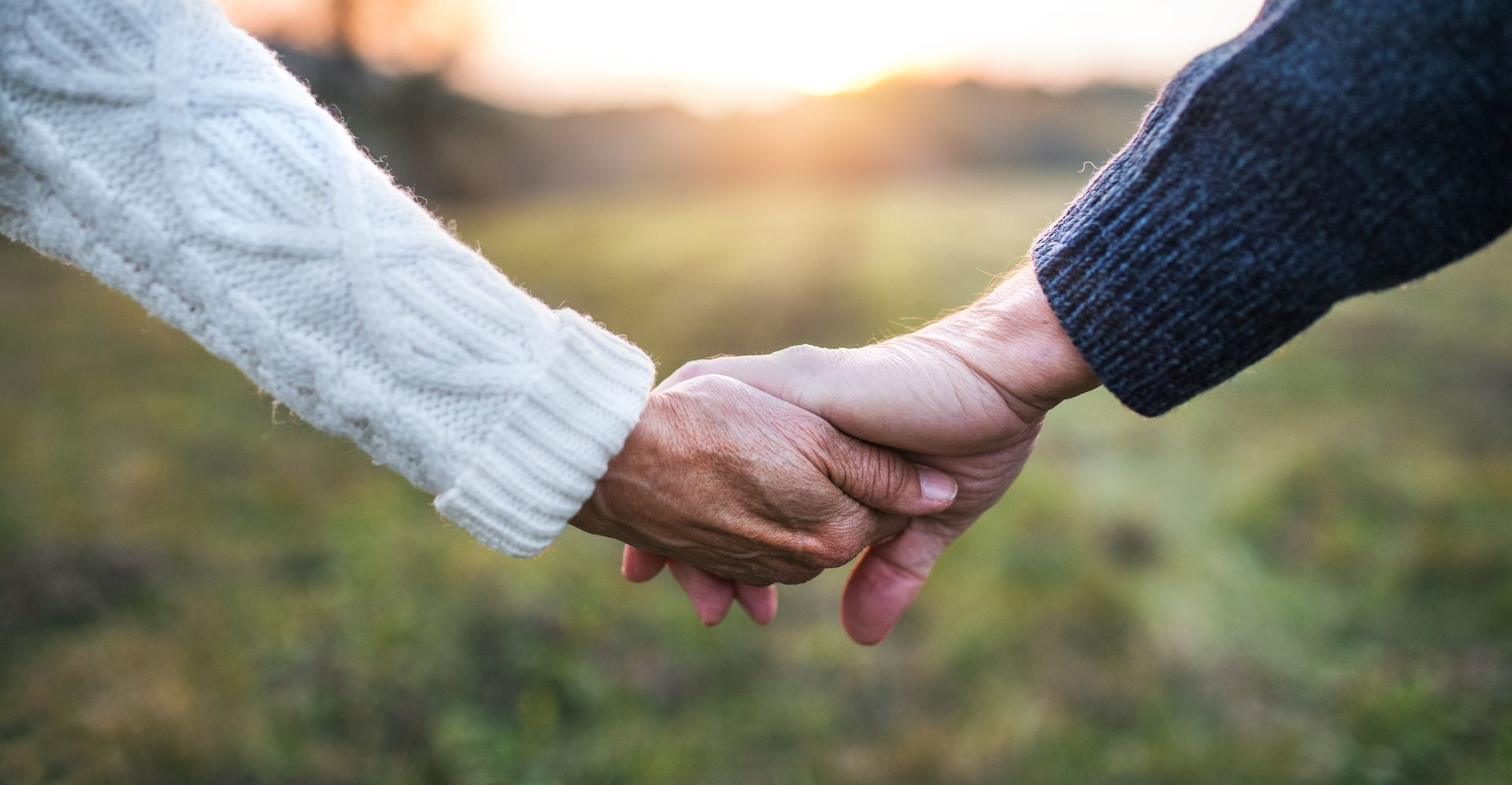 Two people holding hands outdoors at sunset, one in a white sweater and one in a blue sweater.
