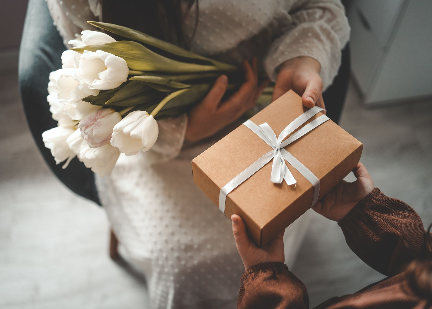 Hands exchanging a small wrapped gift with white tulips in a cozy indoor setting