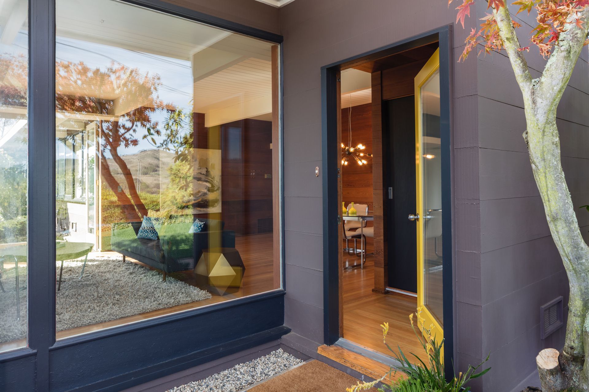 A yellow door stands open to a warm, dimly lit room next to a large floor-to-ceiling window reflecting a garden.