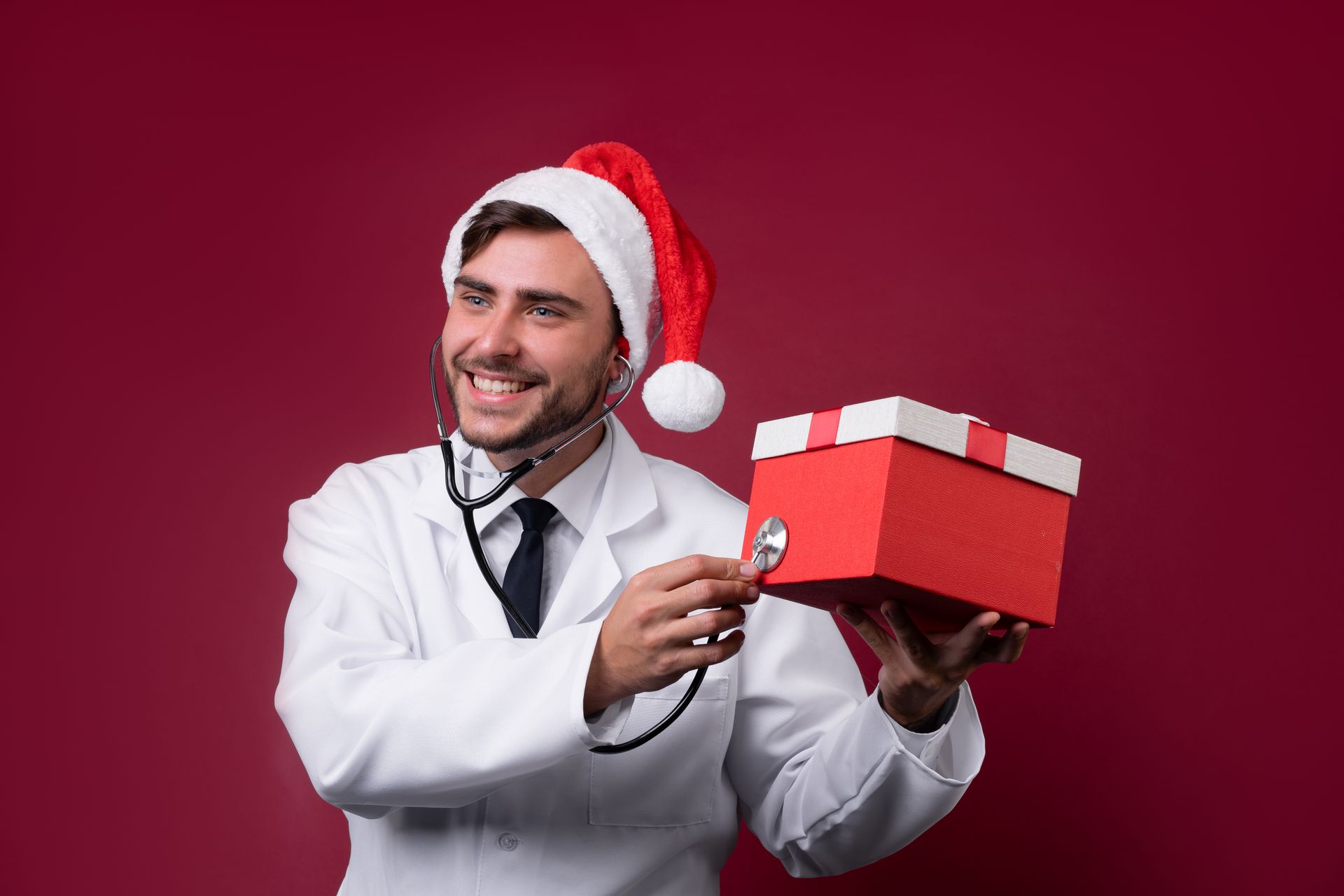 Presente para médicos no natal Médico com gorro de Papai Noel usando um estetoscópio para examinar uma caixa de presente vermelha.