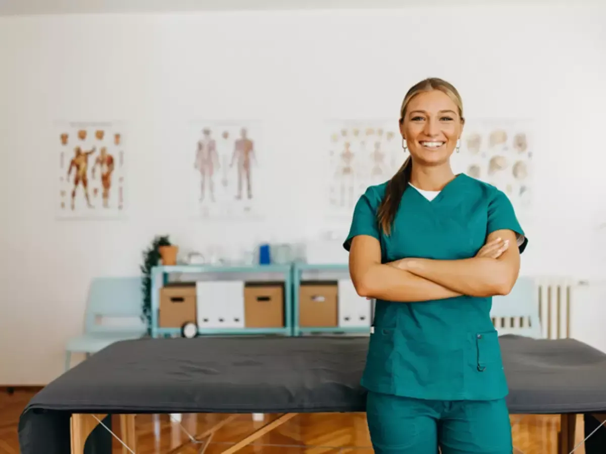 Ficha de anamnese para avaliação fisioterapêutica Uma pessoa sorridente, vestindo um uniforme cirúrgico verde-azulado, está de braços cruzados em um consultório médico, perto de uma maca de massagem.